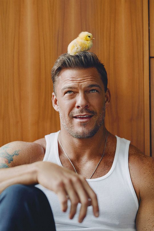 a man with a yellow flower on his head sitting in front of a wooden wall