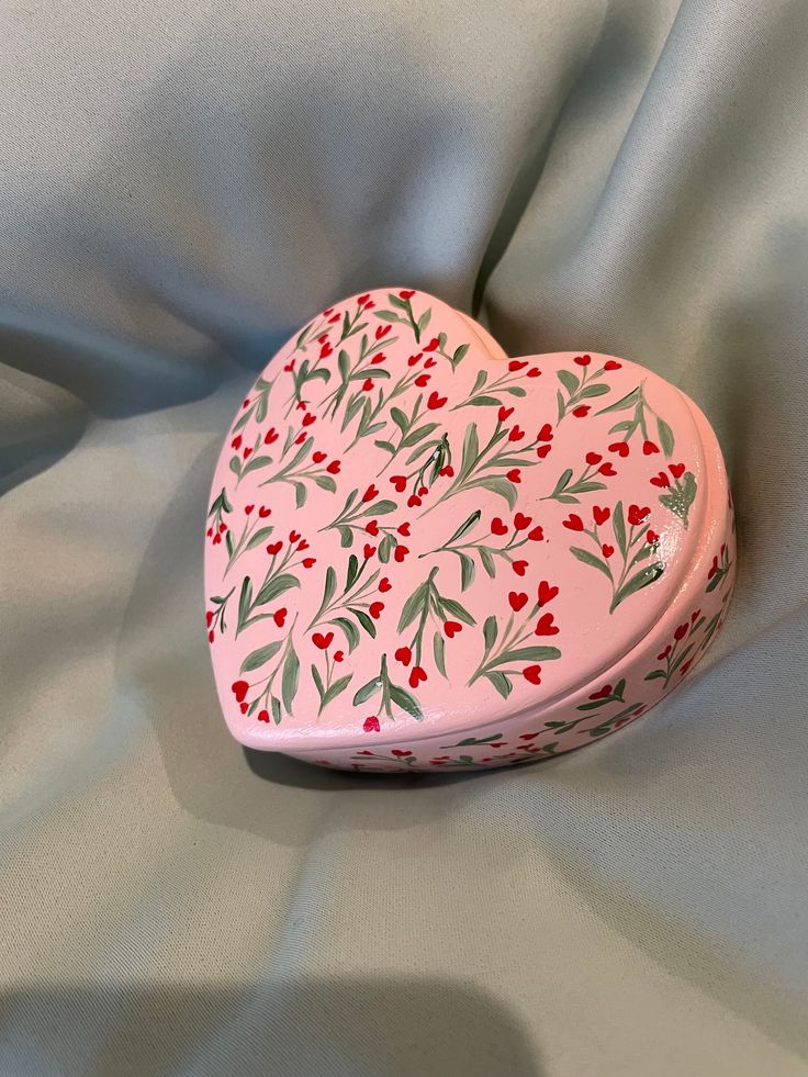 a pink heart shaped box sitting on top of a white cloth covered bedding with red and green leaves