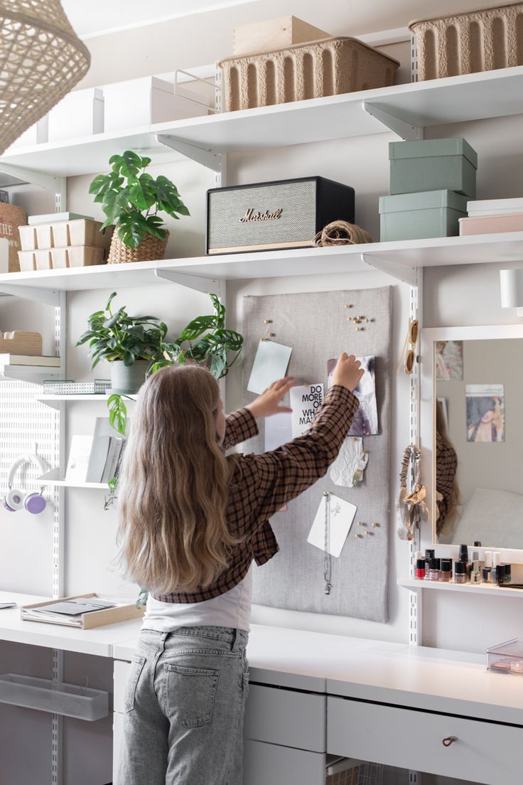 a woman standing in front of a white desk with lots of shelves and plants on it