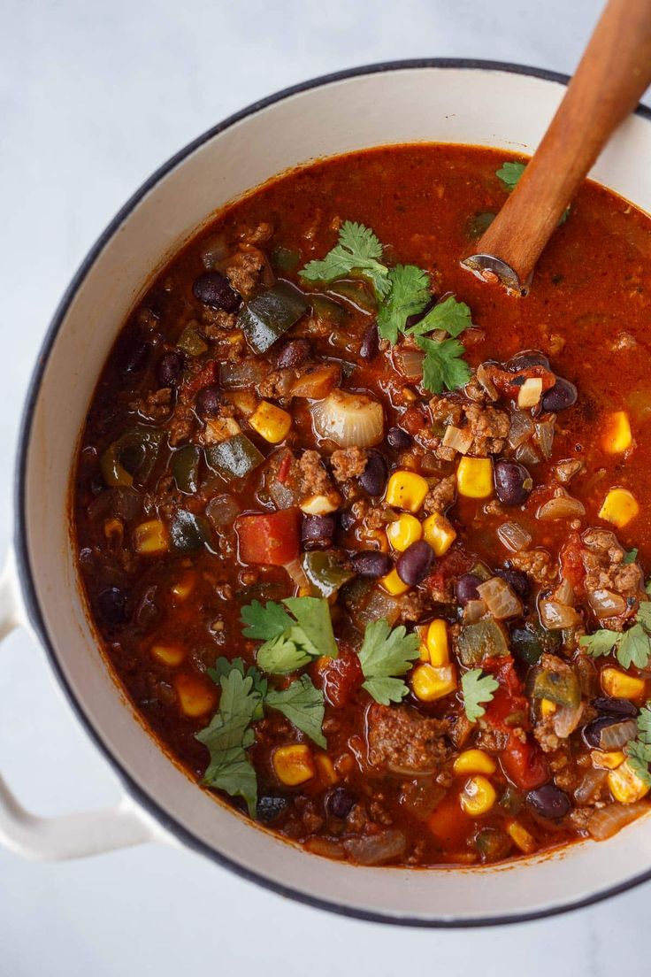 a white bowl filled with chili and corn next to a wooden spoon on top of a table