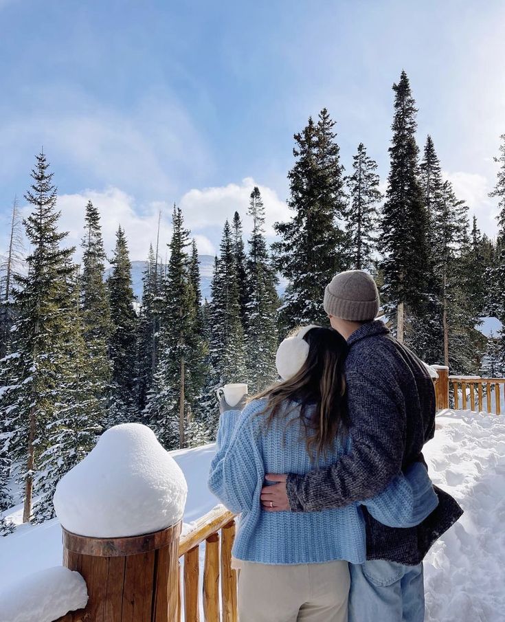 a man and woman standing on top of a snow covered slope next to pine trees