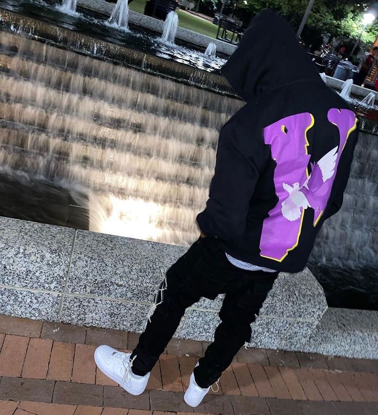 a person wearing a purple and black hoodie is standing near a fountain with water casing in the background