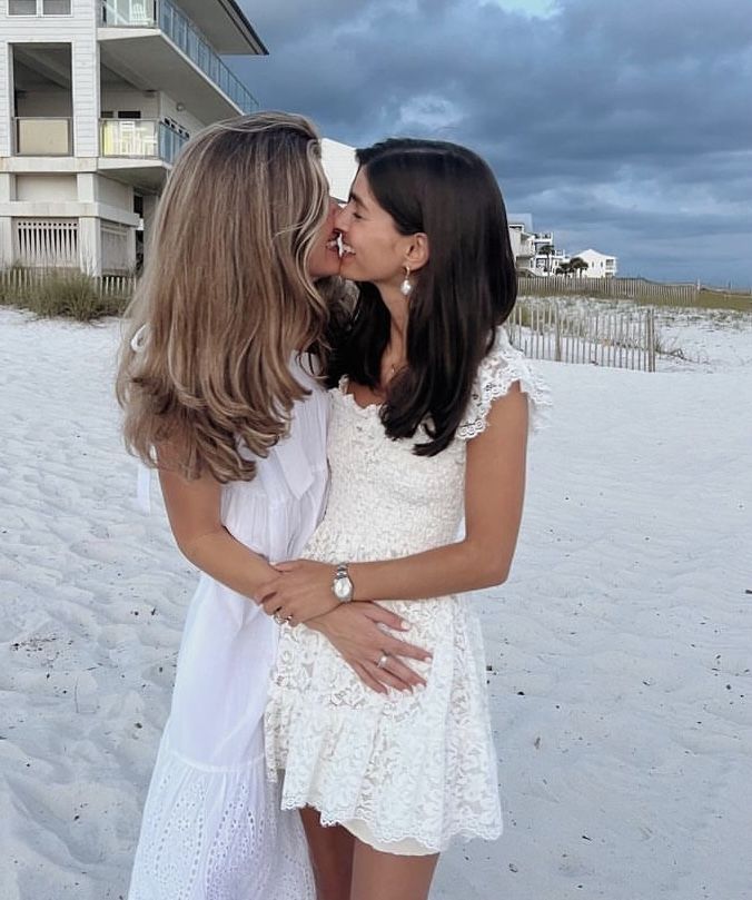 two women kissing on the beach in front of a white house with dark clouds behind them