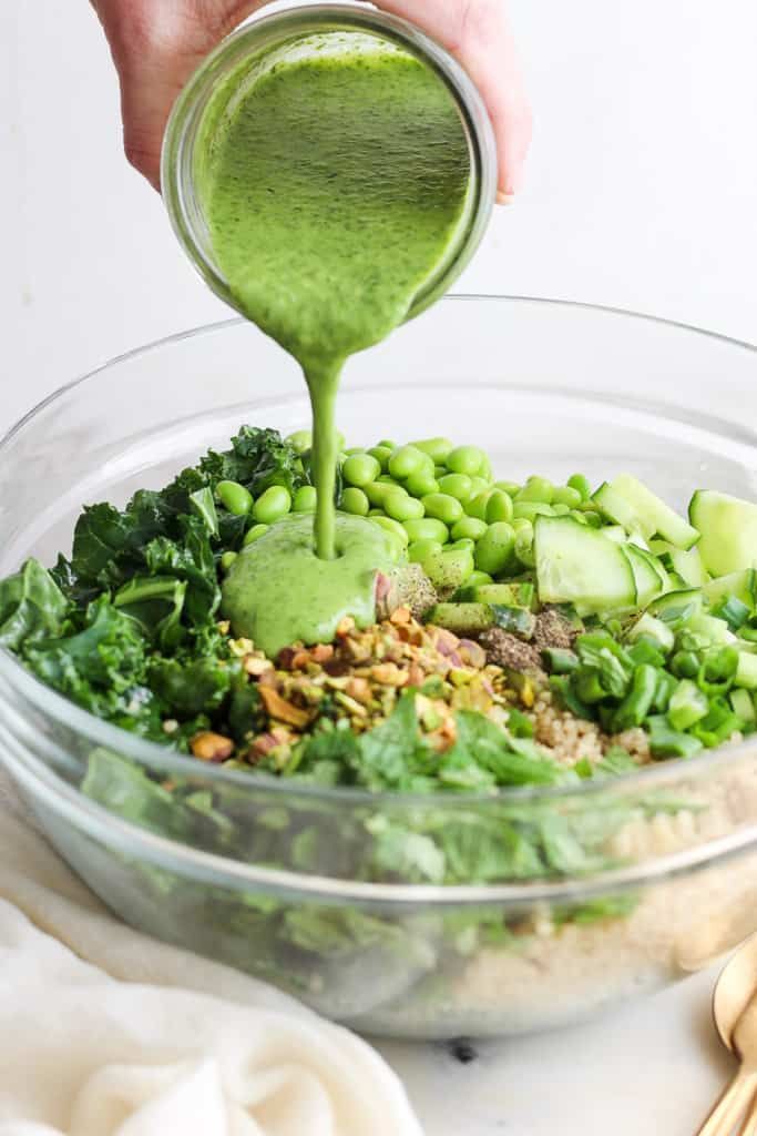 someone pouring green liquid into a bowl filled with vegetables and grains, including broccoli