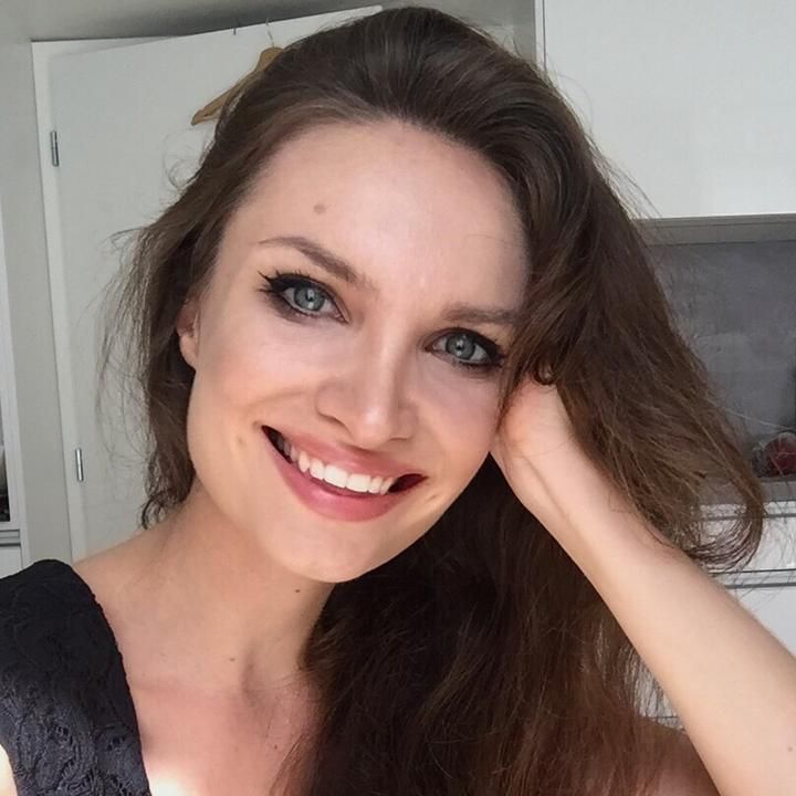 a woman with long brown hair and blue eyes smiles at the camera while holding her hand on her head