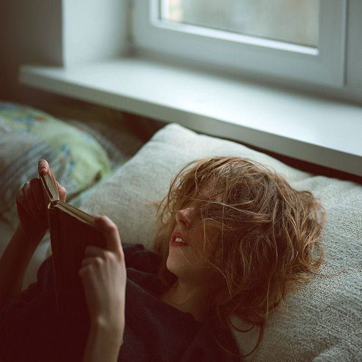 a woman laying on top of a bed next to a windowsill reading a book