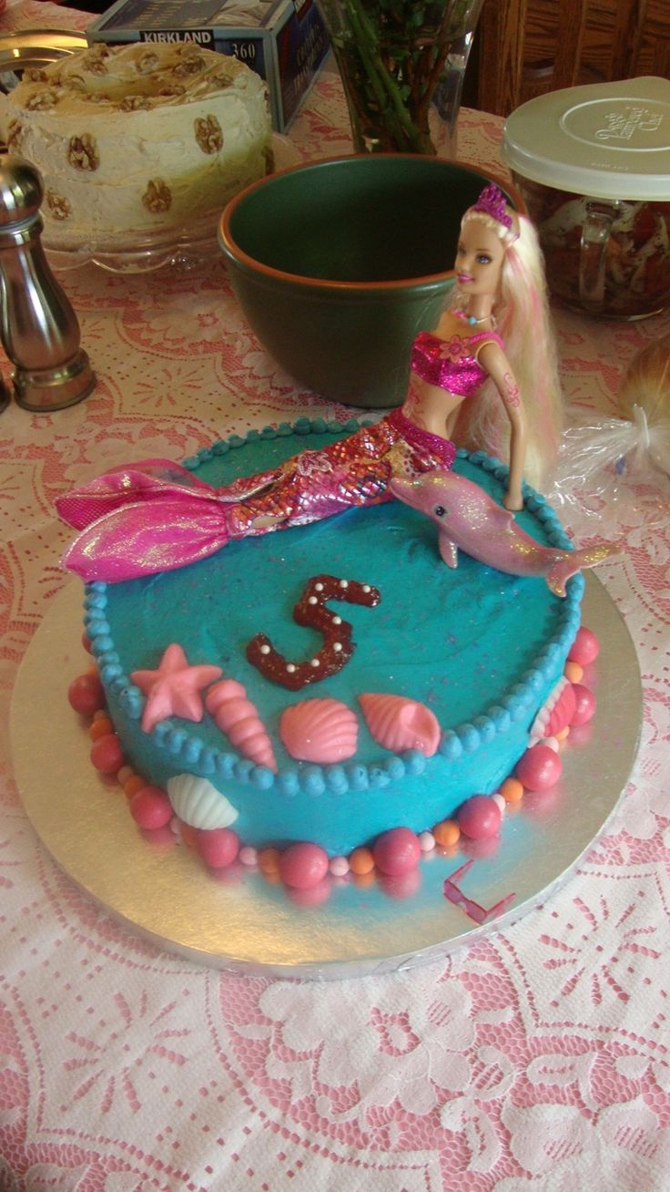 a birthday cake with a mermaid doll on top and decorations around the edges, sitting on a table