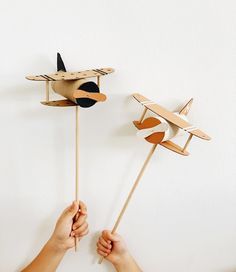 two handmade wooden airplanes on sticks being held by someone's hands against a white wall