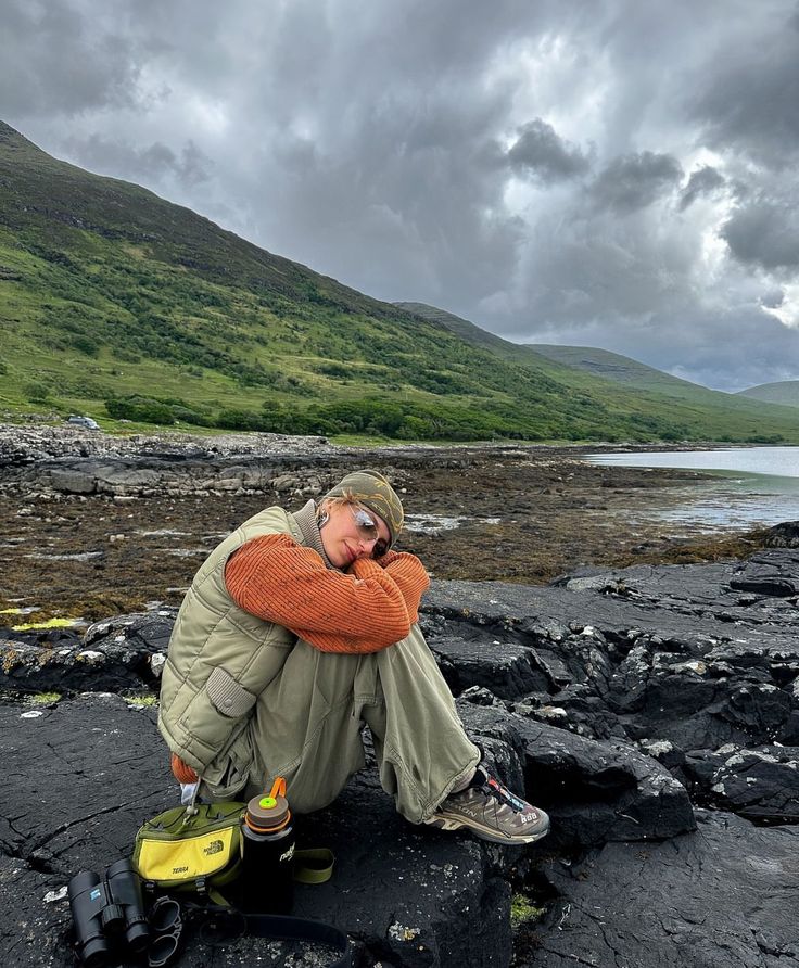 a man sitting on top of a rocky beach next to a body of water with mountains in the background