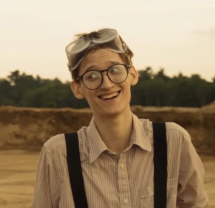 a young man wearing glasses and suspenders smiles at the camera with trees in the background