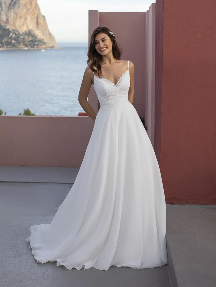 a woman in a white wedding dress standing on a balcony next to the ocean with her hands on her hips