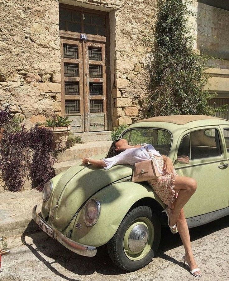 a woman leaning on the hood of an old green car in front of a stone building