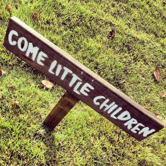 a wooden sign that says come little children on it in the grass next to some leaves