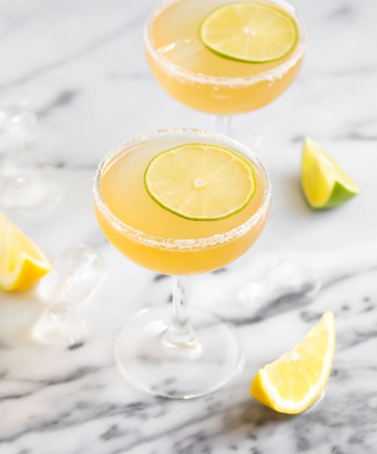 two glasses filled with lemonade and limes on top of a marble countertop