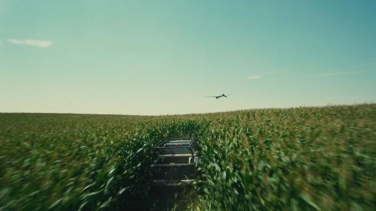 an airplane flying over a corn field with a truck in the foreground and another vehicle on the far side