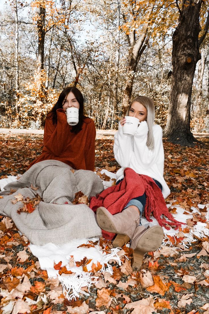 two women are sitting on the ground covered in leaves and drinking from mugs,