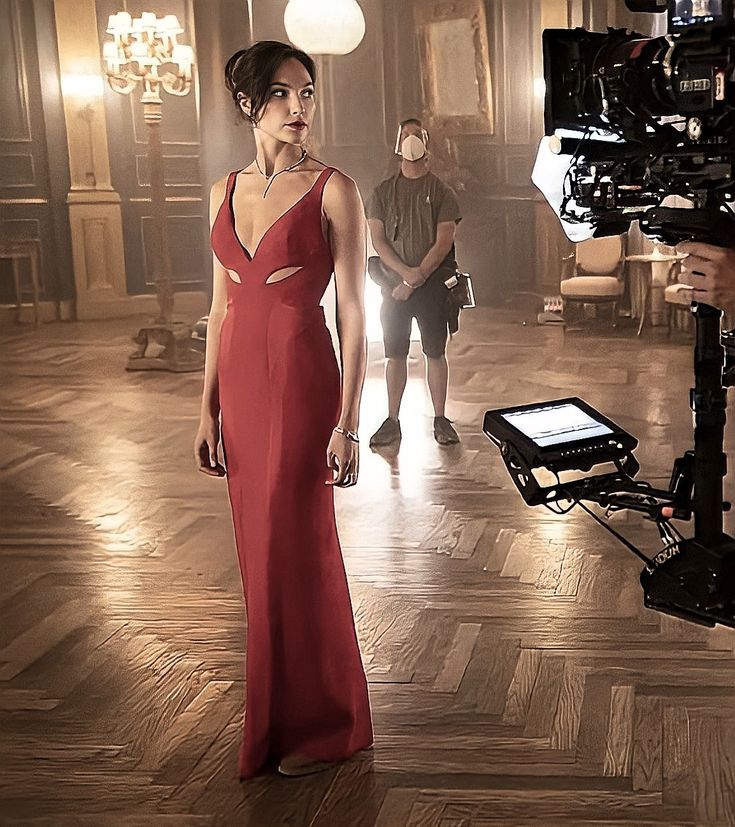 a woman in a long red dress standing next to a camera and another man behind her