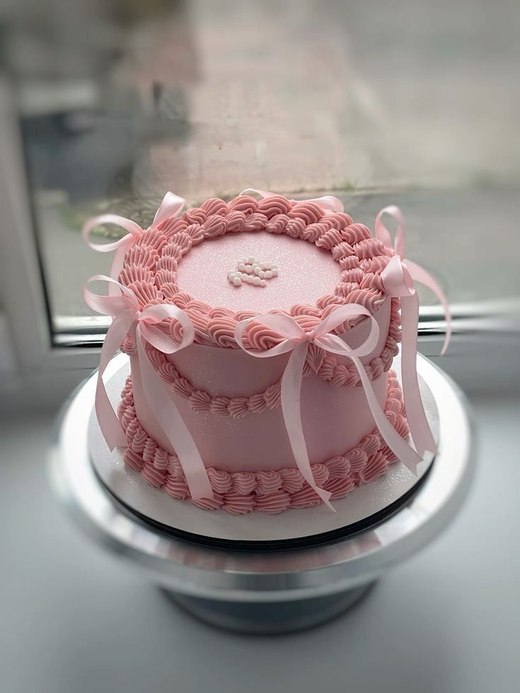 a pink cake sitting on top of a silver plate next to a window with white ribbons