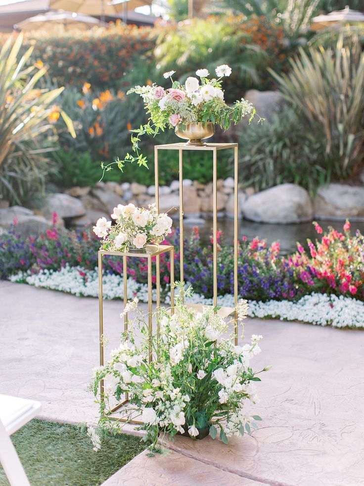 three tiered metal plant stands with flowers on them in front of some rocks and plants