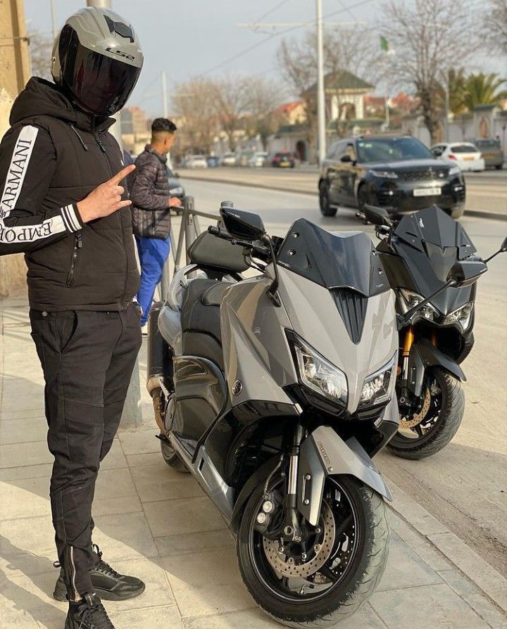 a man standing next to a parked motorcycle on the side of a road with another person looking at his cell phone