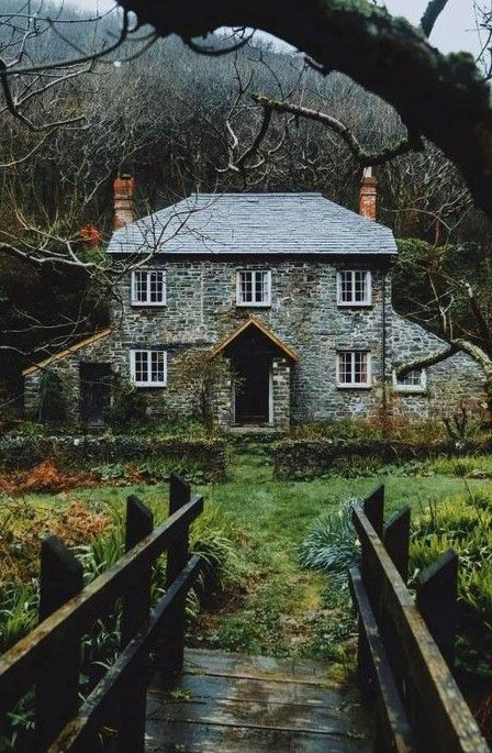 an old stone house sitting in the middle of a lush green field next to a wooden bridge