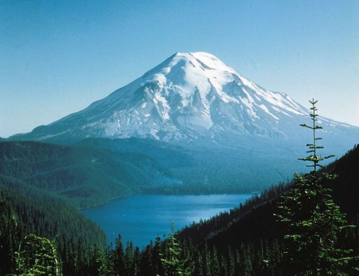 a snow covered mountain towering over a lake surrounded by trees and evergreens in the foreground