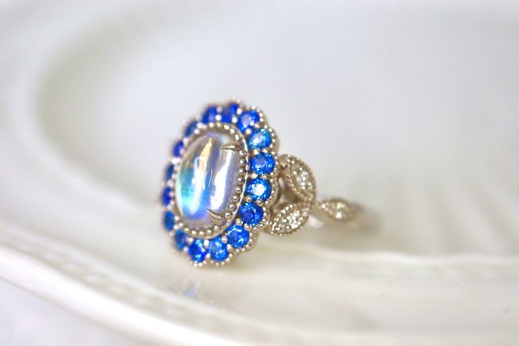 Mesmerizing Blue Moonstone Halo Ring. Moonstone engagement ring. Blue sapphires & diamonds. Edwardian Moonstone. 14K White Gold. US 7.25 A beautiful and unique moonstone ring, created with precision cut sapphires in 14K white gold, a luxurious and high-quality piece of jewelry, perfect to express your love. 💎 PREMIUM RING -- A brand new moonstone ring, personally designed and made with the highest quality of 14K white gold.  ✨ CLASSIC BRILLIANCE -- Designed with a beautiful blue moonstone at th Vintage Blue Sapphire Ring With Halo Setting, Vintage Blue Halo Setting Jewelry, Vintage Blue Jewelry With Halo Setting, Blue Oval Moonstone Ring, Formal Blue Moonstone Gemstone Ring, Unique Blue Moonstone Ring, Unique Blue Sapphire Ring For Wedding, Blue Moonstone Ring With Accent Stones, Sapphire Moonstone Anniversary Ring