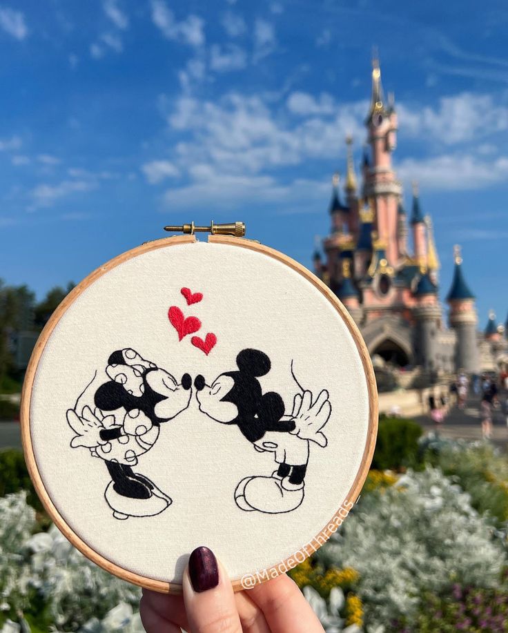 a person holding up a cross stitch mickey and minnie mouse in front of a castle