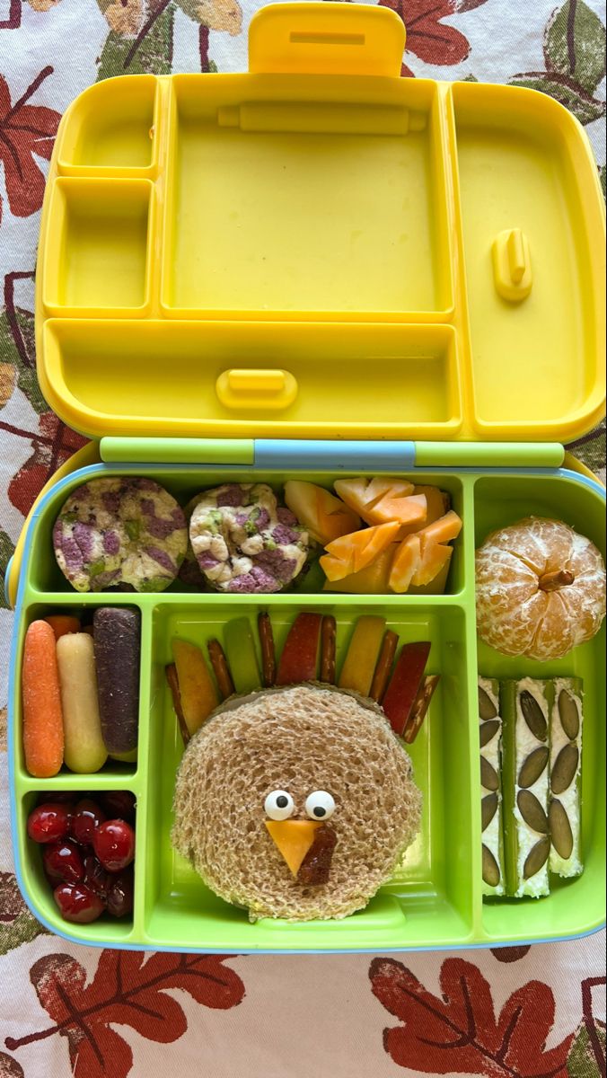 an open lunch box with food in it and a turkey face on the lid, sitting on a bed