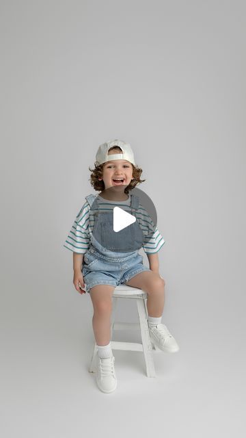 a little boy sitting on top of a stool wearing overalls and a hat with his mouth open