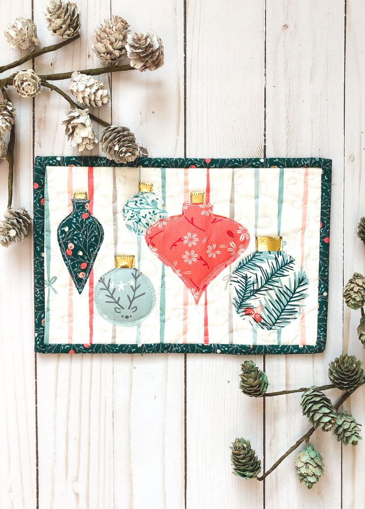 an embroidered christmas ornament hanging on a white wooden wall next to pine cones