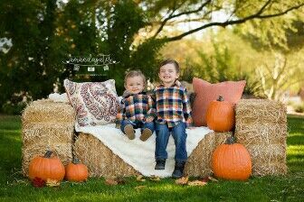 two young boys sitting on hay bales with pumpkins in the grass behind them