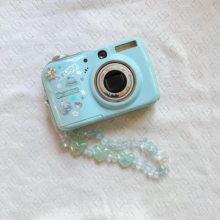 a blue camera sitting on top of a white bed next to a beaded necklace