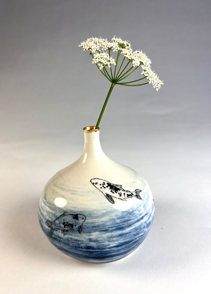 a white and blue vase with a flower in it on a gray background, the bottom half has a fish drawn on it