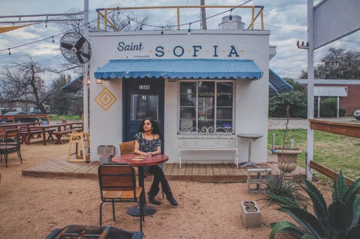 a woman sitting at a table in front of a small building with a blue awning