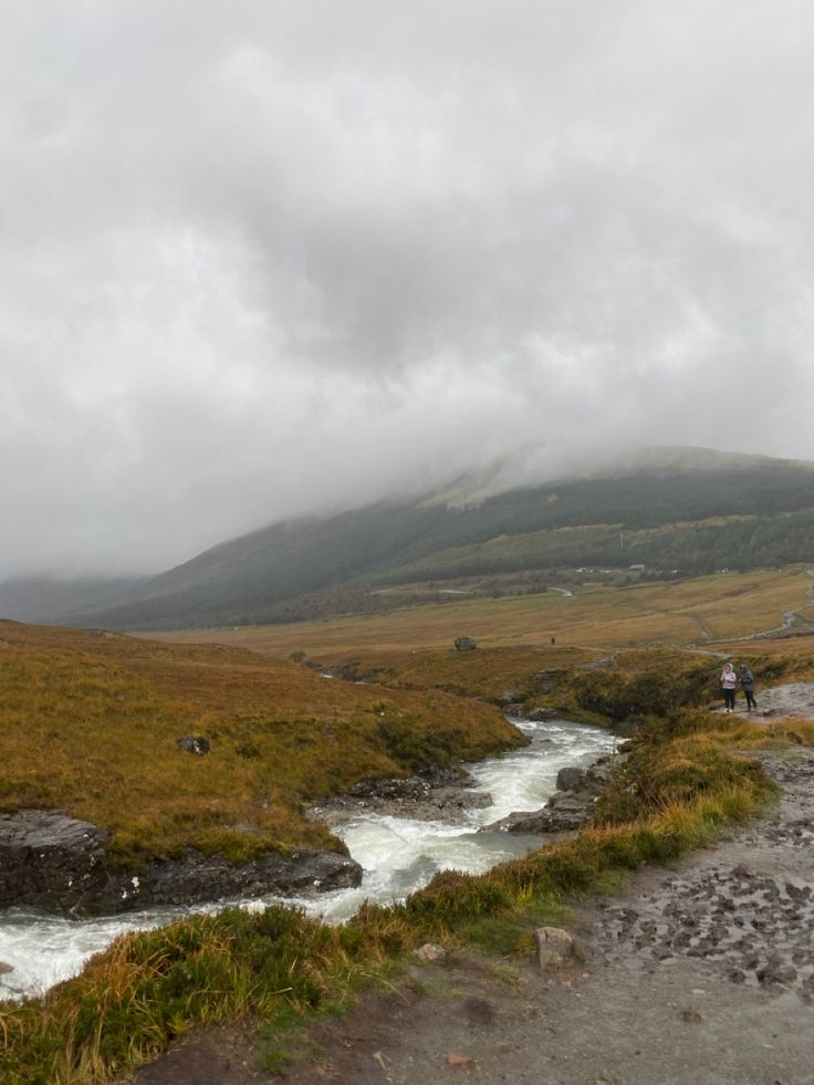 two people are standing on the side of a mountain with a river running through it