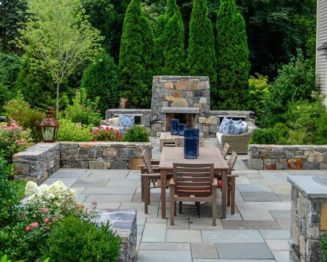 an outdoor dining area with stone walls and seating around the fire place, surrounded by greenery