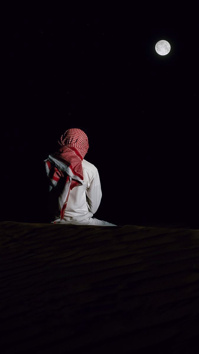 a man sitting on top of a sand dune under a moon lit sky with a red and white scarf