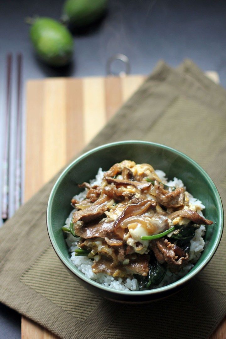 a green bowl filled with rice and meat on top of a wooden cutting board next to silverware