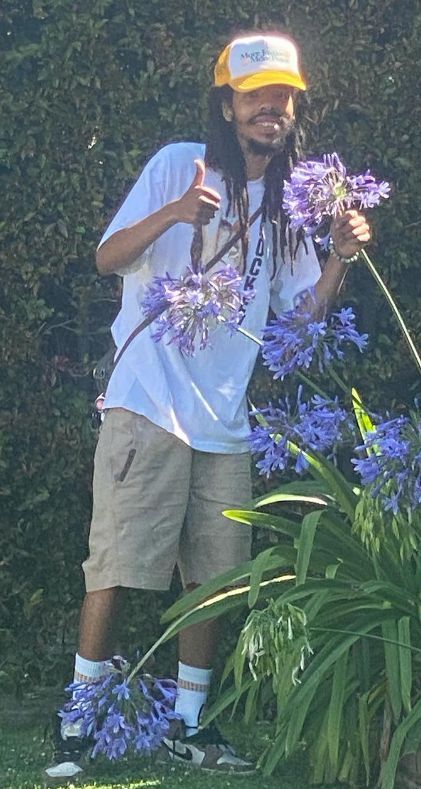 a man with dreadlocks standing in front of purple flowers and holding a yellow hat