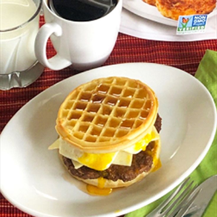 a waffle sandwich on a white plate next to a cup of coffee and milk