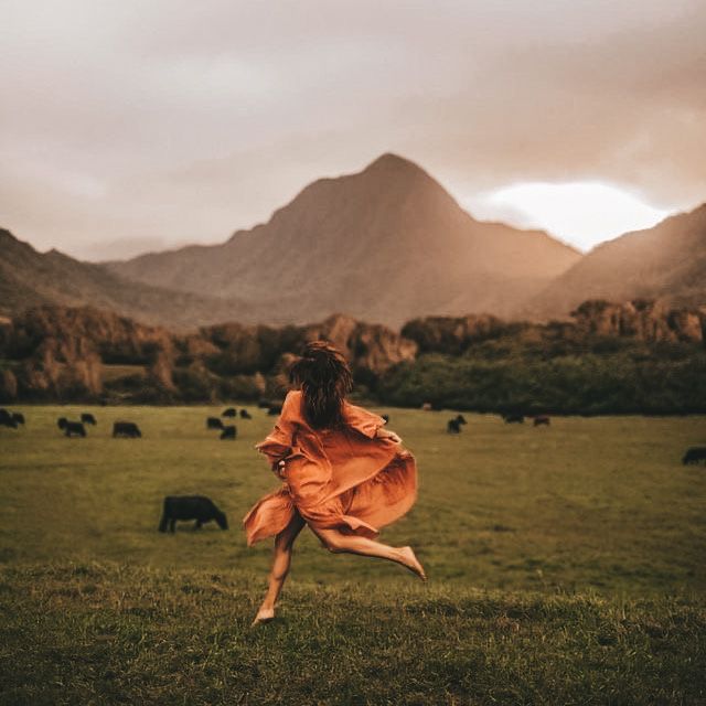 a woman in an orange dress is running through a field with cows and mountains behind her