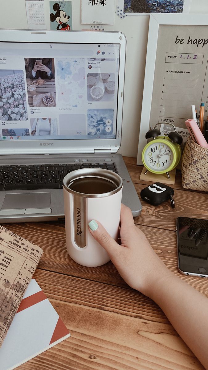 a person holding a coffee cup in front of a laptop on a desk with other items