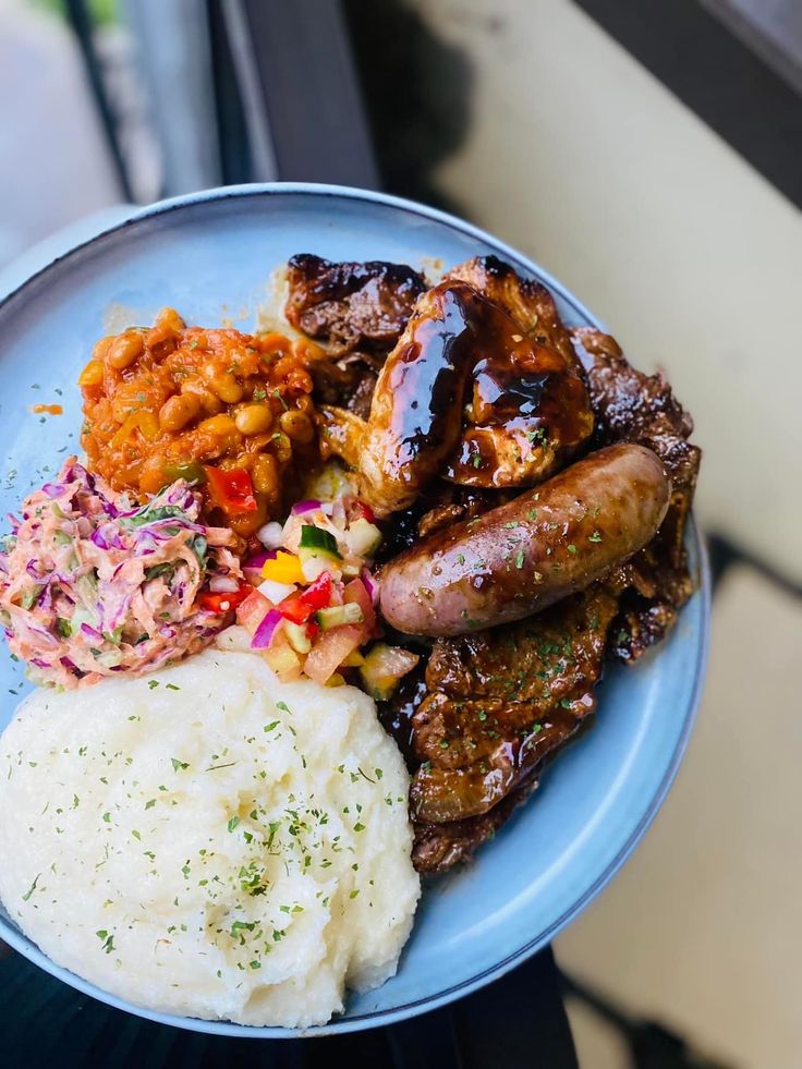 a blue plate topped with meat, mashed potatoes and veggies next to a window