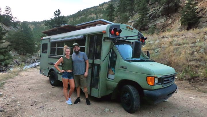 two people standing in front of a green bus on the side of a dirt road