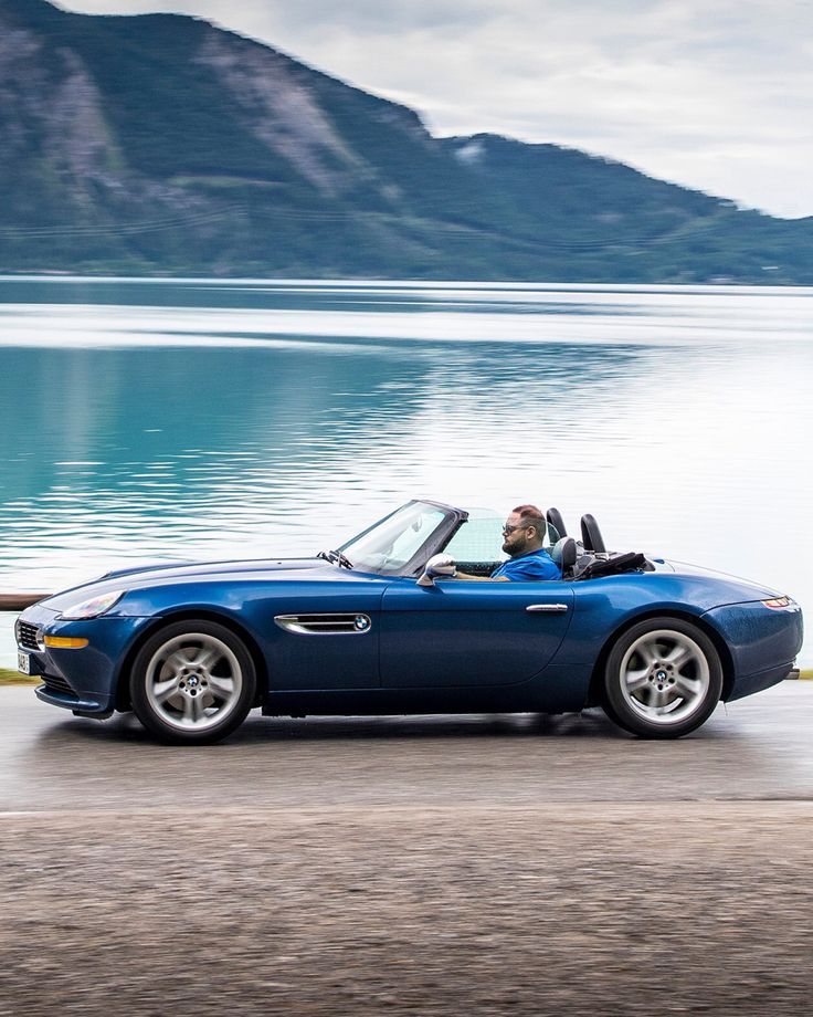 a blue sports car driving down the road next to a lake with mountains in the background