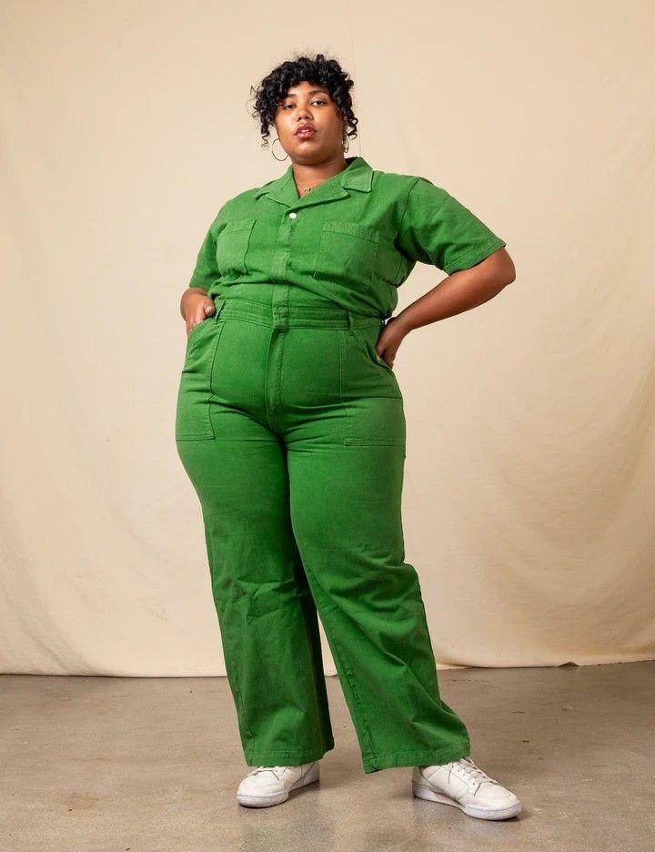 a woman standing in front of a white backdrop wearing green overalls and white tennis shoes