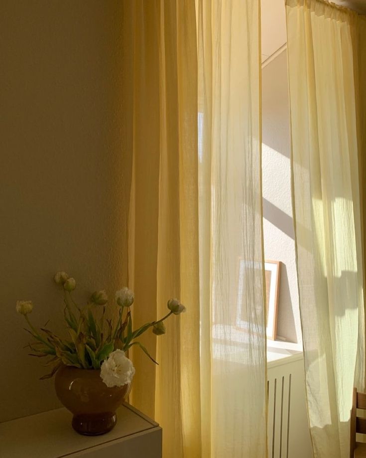 a vase with white flowers sitting on top of a table next to a window covered in sheer curtains