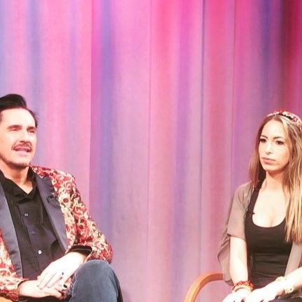 a man and woman sitting on chairs in front of a colorful curtain, talking to each other