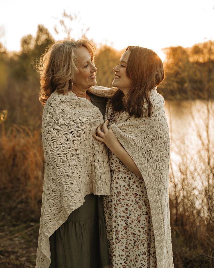 two women standing next to each other in front of a body of water wearing shawls